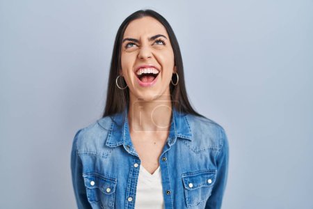 Photo for Hispanic woman standing over blue background angry and mad screaming frustrated and furious, shouting with anger. rage and aggressive concept. - Royalty Free Image
