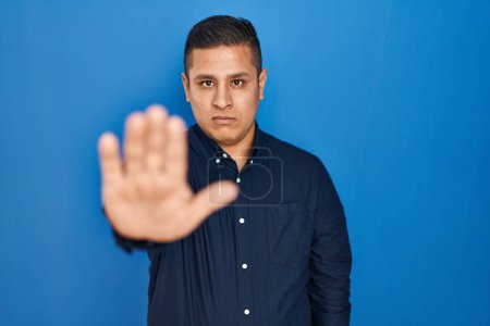Photo for Hispanic young man standing over blue background doing stop sing with palm of the hand. warning expression with negative and serious gesture on the face. - Royalty Free Image