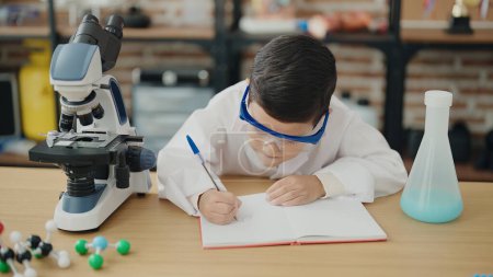 Photo for Adorable hispanic boy student using microscope writing on notebook at laboratory classroom - Royalty Free Image