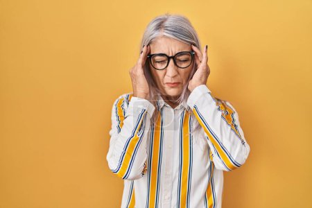Téléchargez les photos : Femme d'âge moyen avec les cheveux gris debout sur fond jaune portant des lunettes avec la main sur la tête pour la douleur à la tête parce que le stress. migraine souffrant. - en image libre de droit