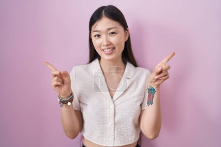 Foto de Mujer joven china de pie sobre fondo rosa sonriendo confiado señalando con los dedos a diferentes direcciones. espacio de copia para publicidad - Imagen libre de derechos