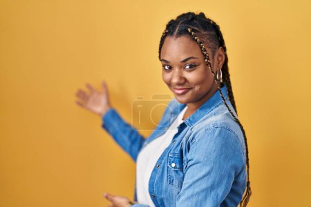 Photo for African american woman with braids standing over yellow background inviting to enter smiling natural with open hand - Royalty Free Image