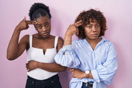 Téléchargez les photos : Deux femmes africaines debout sur fond rose tirant et se suicidant en pointant la main et les doigts vers la tête comme une arme à feu, geste de suicide. - en image libre de droit