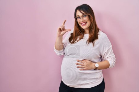 Téléchargez les photos : Pregnant woman standing over pink background smiling and confident gesturing with hand doing small size sign with fingers looking and the camera. measure concept. - en image libre de droit