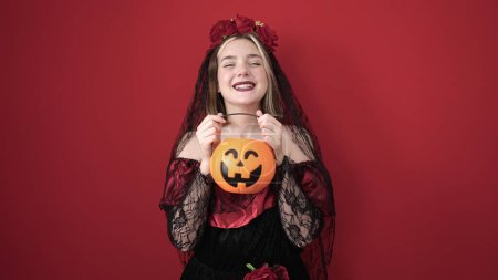Foto de Young blonde woman wearing katrina costume holding halloween pumpkin basket over isolated red background - Imagen libre de derechos