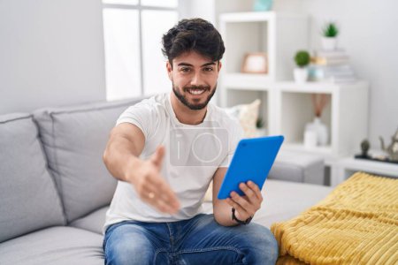Foto de Hispanic man with beard using touchpad sitting on the sofa smiling friendly offering handshake as greeting and welcoming. successful business. - Imagen libre de derechos
