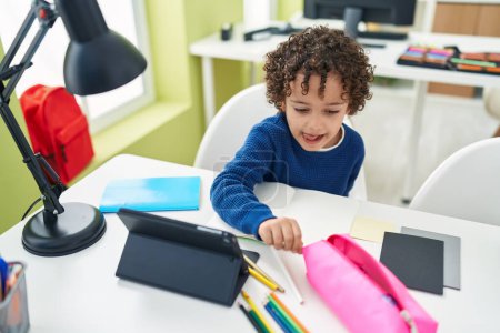 Photo for Adorable hispanic boy preschool student sitting on table drawing on notebook at classroom - Royalty Free Image