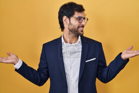 Téléchargez les photos : Handsome latin man standing over yellow background smiling showing both hands open palms, presenting and advertising comparison and balance - en image libre de droit