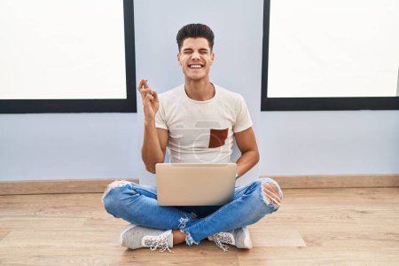 Foto de Young hispanic man using laptop at home gesturing finger crossed smiling with hope and eyes closed. luck and superstitious concept. - Imagen libre de derechos