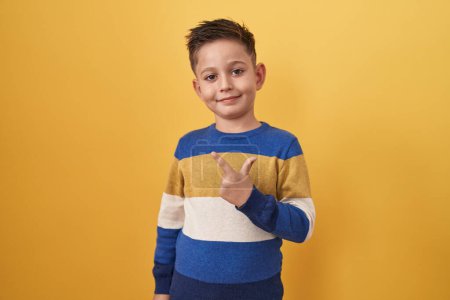 Foto de Pequeño niño hispano de pie sobre fondo amarillo sonriendo alegre señalando con la mano y el dedo hacia un lado - Imagen libre de derechos