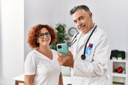 Téléchargez les photos : Homme et femme d'âge moyen portant l'uniforme de médecin utilisant le smartphone ayant la consultation médicale à la clinique - en image libre de droit