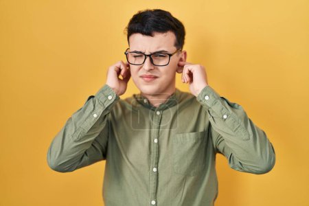 Téléchargez les photos : Non binary person standing over yellow background covering ears with fingers with annoyed expression for the noise of loud music. deaf concept. - en image libre de droit