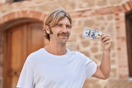 Téléchargez les photos : Young blond man smiling confident holding 100 dollars banknote at street - en image libre de droit