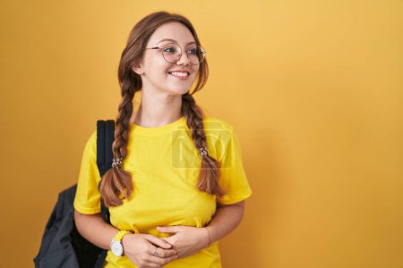 Photo for Young caucasian woman wearing student backpack over yellow background looking away to side with smile on face, natural expression. laughing confident. - Royalty Free Image