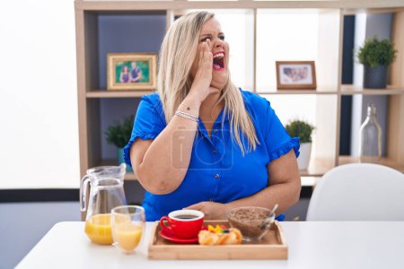 Téléchargez les photos : Caucasian plus size woman eating breakfast at home shouting and screaming loud to side with hand on mouth. communication concept. - en image libre de droit