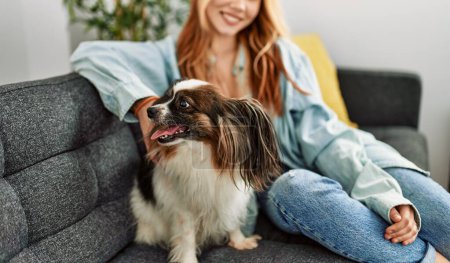 Foto de Joven mujer caucásica sonriendo confiado sentado en sofá con perro en casa - Imagen libre de derechos