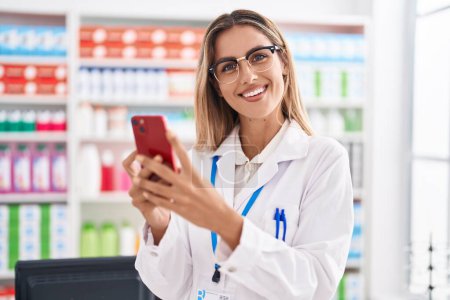 Téléchargez les photos : Young blonde woman pharmacist using smartphone working at pharmacy - en image libre de droit