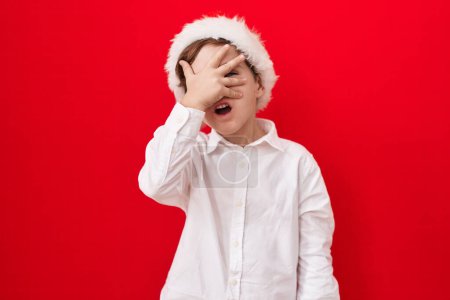 Foto de Pequeño niño caucásico con sombrero de navidad sobre fondo rojo asomándose en shock cubriendo la cara y los ojos con la mano, mirando a través de los dedos con miedo - Imagen libre de derechos