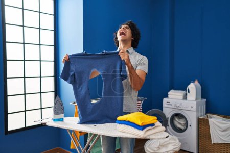 Foto de Hombre hispano con pelo rizado planchando sosteniendo camisa de hierro quemada en la lavandería enojado y loco gritando frustrado y furioso, gritando con ira mirando hacia arriba. - Imagen libre de derechos