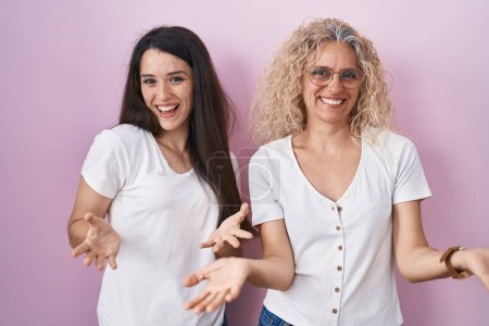 Téléchargez les photos : Mère et fille debout ensemble sur fond rose souriant gai avec les bras ouverts comme accueil amical, salutations positives et confiantes - en image libre de droit
