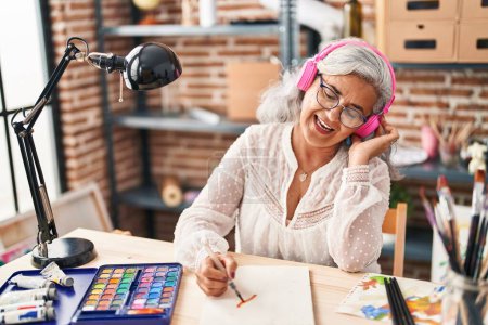 Photo for Middle age woman artist drawing on notebook listening to music at art studio - Royalty Free Image