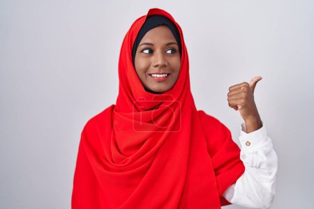 Téléchargez les photos : Jeune femme arabe portant l'écharpe traditionnelle islamique hijab souriant avec le visage heureux regardant et pointant vers le côté avec le pouce vers le haut. - en image libre de droit