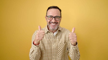 Téléchargez les photos : Moyen Age homme souriant confiant faire ok signe avec les pouces vers le haut sur fond jaune isolé - en image libre de droit