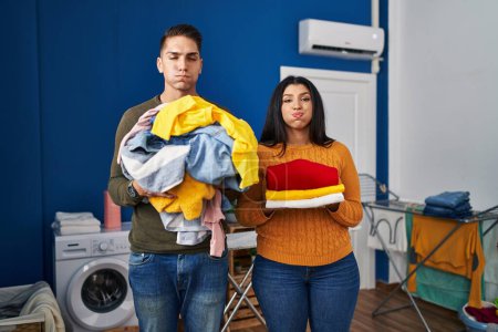 Foto de Young couple holding laundry dirty and clean laundry puffing cheeks with funny face. mouth inflated with air, catching air. - Imagen libre de derechos