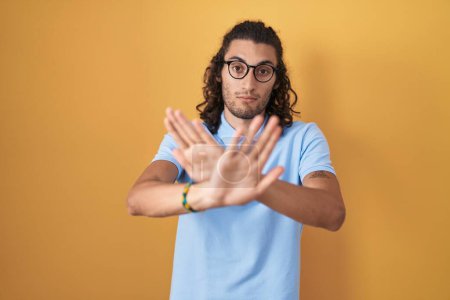 Photo for Young hispanic man standing over yellow background rejection expression crossing arms and palms doing negative sign, angry face - Royalty Free Image
