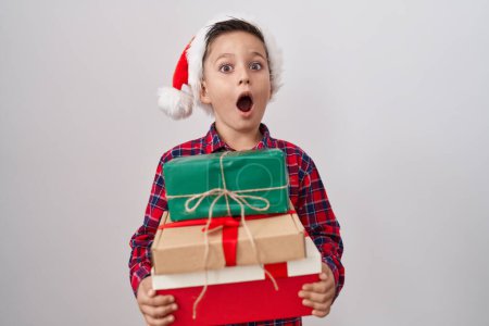 Téléchargez les photos : Little hispanic boy wearing christmas hat holding presents afraid and shocked with surprise and amazed expression, fear and excited face. - en image libre de droit