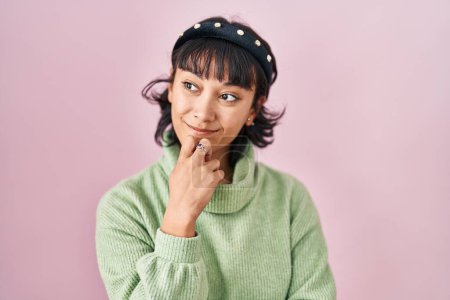 Foto de Young beautiful woman standing over pink background with hand on chin thinking about question, pensive expression. smiling and thoughtful face. doubt concept. - Imagen libre de derechos