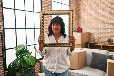 Foto de Hispanic woman at new home holding empty frame clueless and confused expression. doubt concept. - Imagen libre de derechos