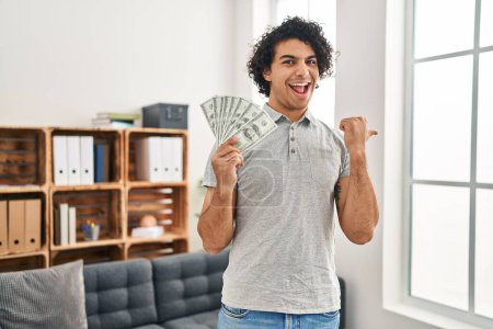 Photo for Hispanic man with curly hair holding 100 dollars banknotes pointing thumb up to the side smiling happy with open mouth - Royalty Free Image