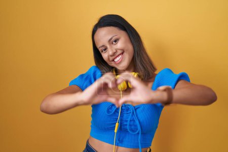 Téléchargez les photos : Jeune femme hispanique debout sur fond jaune souriant dans l'amour faisant forme de symbole de coeur avec les mains. concept romantique. - en image libre de droit