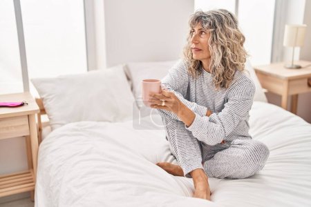 Foto de Mujer de mediana edad bebiendo taza de café sentado en la cama en el dormitorio - Imagen libre de derechos
