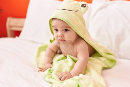 Foto de Adorable niño hispano usando una toalla divertida sentado en la cama en el dormitorio - Imagen libre de derechos