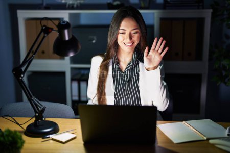Téléchargez les photos : Jeune femme brune travaillant au bureau la nuit avec un ordinateur portable renonçant à dire bonjour heureux et souriant, geste de bienvenue amical - en image libre de droit