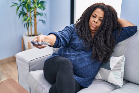Foto de African american woman watching tv lying on sofa with boring expression at home - Imagen libre de derechos