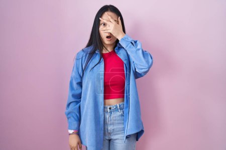 Téléchargez les photos : Young asian woman standing over pink background peeking in shock covering face and eyes with hand, looking through fingers with embarrassed expression. - en image libre de droit