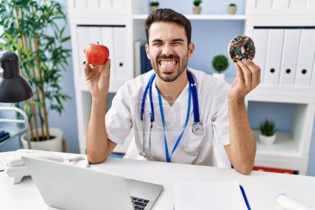 Foto de Joven dietista hispano sosteniendo rosquilla y manzana sacando la lengua feliz con expresión divertida. - Imagen libre de derechos
