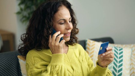 Téléchargez les photos : Moyen Age femme hispanique parler sur smartphone tenant carte de crédit à la maison - en image libre de droit