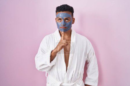 Téléchargez les photos : Young hispanic man wearing beauty face mask and bath robe thinking concentrated about doubt with finger on chin and looking up wondering - en image libre de droit
