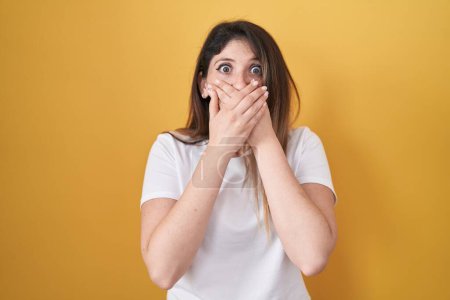 Photo for Young brunette woman standing over yellow background shocked covering mouth with hands for mistake. secret concept. - Royalty Free Image