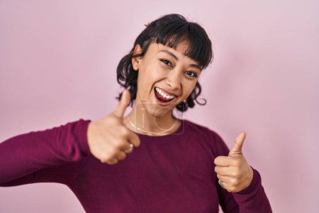 Foto de Young beautiful woman standing over pink background approving doing positive gesture with hand, thumbs up smiling and happy for success. winner gesture. - Imagen libre de derechos