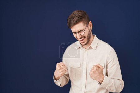 Foto de Hombre hispano con barba de pie sobre fondo azul muy feliz y emocionado haciendo gesto ganador con los brazos levantados, sonriendo y gritando por el éxito. concepto de celebración. - Imagen libre de derechos