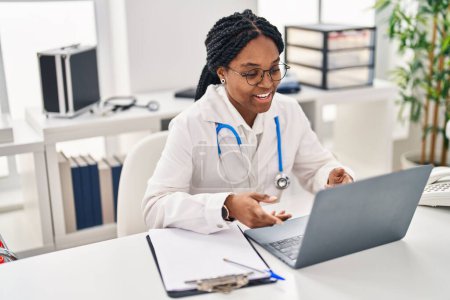 Téléchargez les photos : Femme afro-américaine médecin souriant confiant ayant appel vidéo à la clinique - en image libre de droit