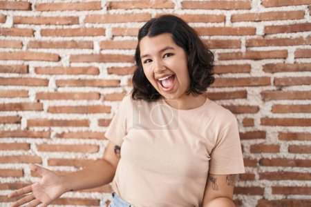 Foto de Mujer hispana joven de pie sobre la pared de ladrillos sonriendo alegre con los brazos abiertos como bienvenida amistosa, saludos positivos y confiados - Imagen libre de derechos