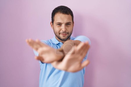 Foto de Hispanic man standing over pink background rejection expression crossing arms and palms doing negative sign, angry face - Imagen libre de derechos