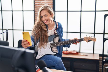 Téléchargez les photos : Jeune femme blonde musicienne jouant de la guitare électrique faire selfie par smartphone au studio de musique - en image libre de droit