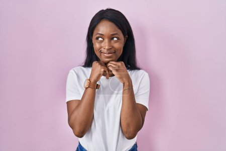 Foto de Mujer joven africana vistiendo camiseta blanca casual riendo nerviosa y emocionada con las manos en la barbilla mirando a un lado - Imagen libre de derechos